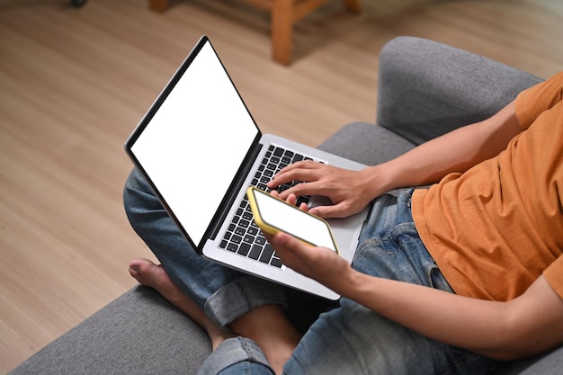 Обрезанный снимок человека, сидящего на диване и использующего ноутбук и смартфон