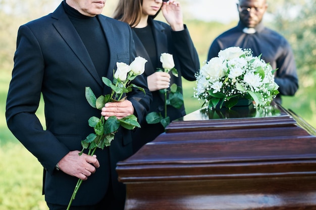 Cropped shot of mature man with two white roses standing by coffin