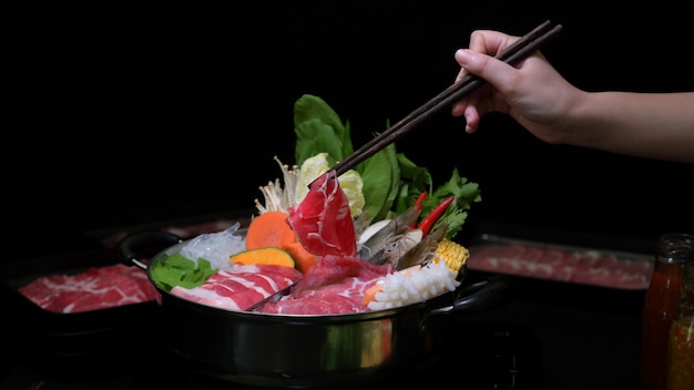 新鮮なスライスした肉、魚介類、野菜と黒の背景を持つ鍋でしゃぶしゃぶを食べる女性のショットをトリミング
