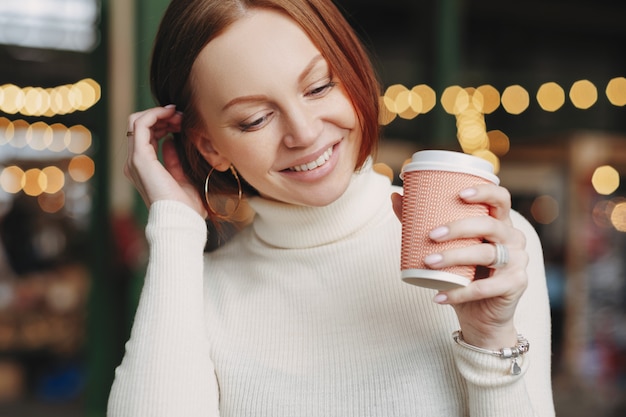 Cropped shot of pleasant looking woman poses in outdoor cafe, holds paper cup of coffee