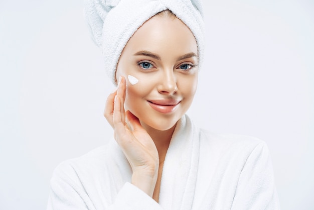 Cropped shot of pretty young woman applies cream for rejuvenation, healthy soft skin, uses cosmetic product, demonstrates nice effect of body lotion, wears comfortable soft white bath robe, towel