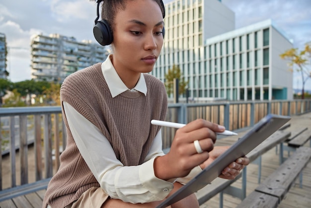 Cropped shot of serious creative woman has inspiration makes drawings on touchpad with stylus listens favorite music via headphones dressed in stylish clothes sits outdoors against urban background