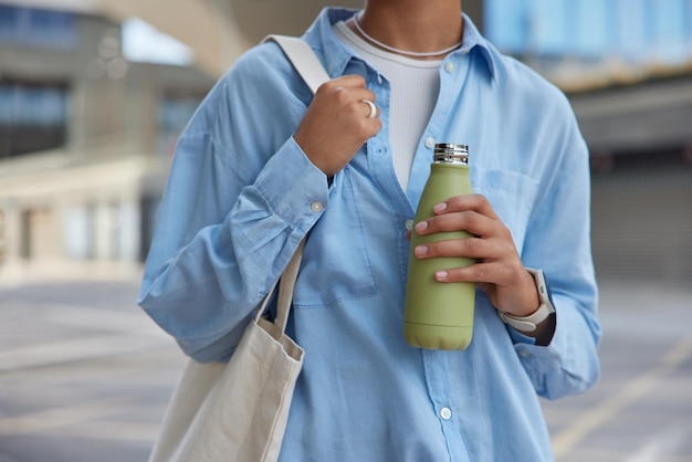 Photo cropped shot of unknown faceless woman wears blue shirt carries fabric bag holds bottle with fresh water strolls at street against blurred background feels thirsty poses against urban setting