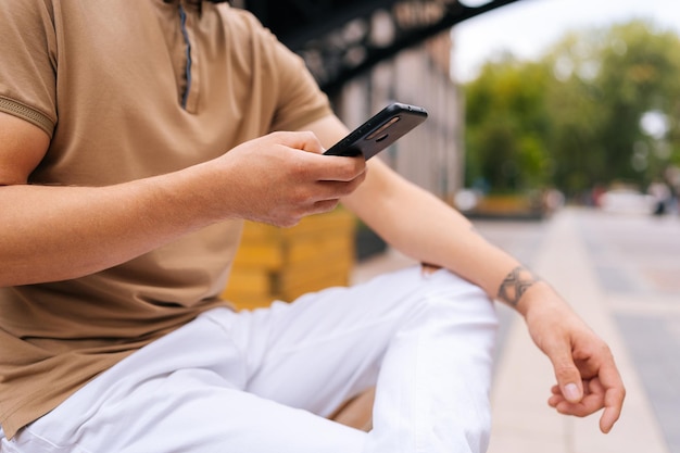 Cropped shot of unrecognizable tattooed male sitting on bench and using typing smartphone looking to screen device on city street