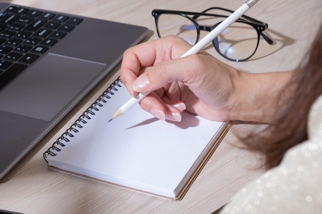 Cropped shot of woman hand writing on notebook with pencil while working with computer at her office desk