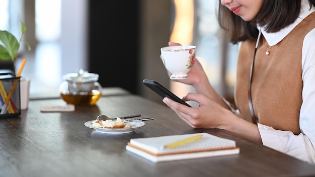 Cropped shot of woman holding a cup of tea and using mobile phone while sitting in cafe