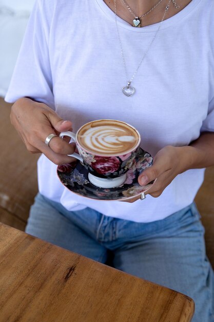 Cropped shot of Woman in white shirt holding cup of hot cappuccino on wooden table top view