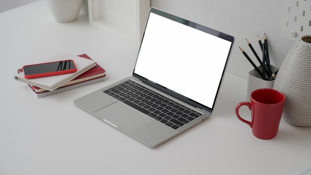 Photo cropped shot of workspace with blank screen laptop, office supplies, decorations and red coffee cup on white desk