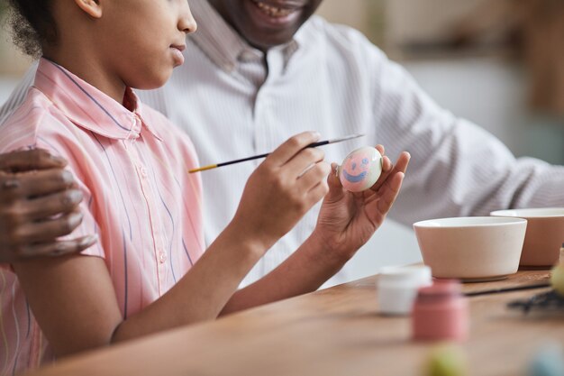Scatto ritagliato di una giovane ragazza afro-americana che dipinge uova di pasqua rosa mentre si gode la decorazione fai-da-te con il padre, copia spazio
