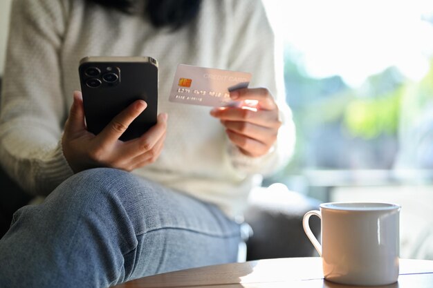 Cropped shot young asian woman holding her mobile phone and\
credit card while sitting in the cafe