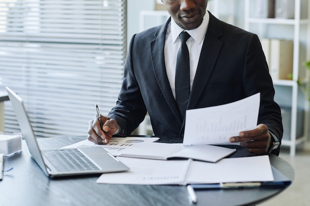cropped shot of young black businessman wearing suit analyzing data reports at workplace in office c
