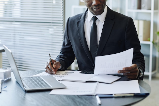 Cropped shot of young black businessman wearing suit analyzing data reports at workplace in office c