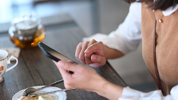 Cropped shot of young female eating dessert and using smart phone while sitting in cafe.