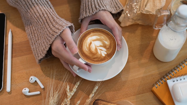 Cropped shot of young female holding hot latte cup on wooden table on working table