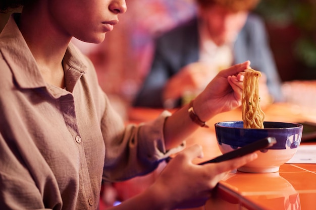 Cropped shot of young hungry woman with cellphone having ramen