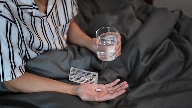 Cropped shot young man holding pill and glass of water while sitting on bed