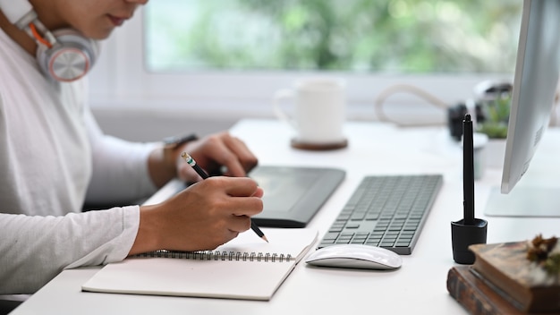 Cropped shot of young man sitting in front of computer and writing important ideas in a notebook.