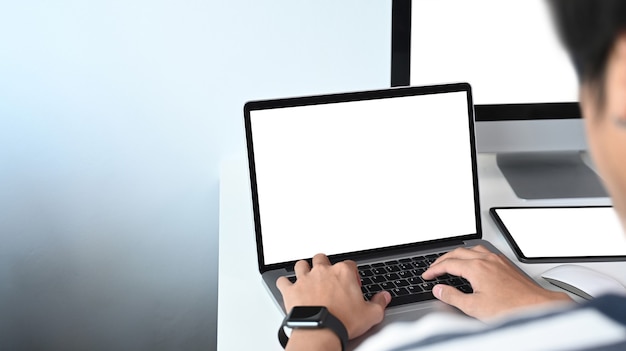 Обрезанный снимок молодого человека, работающего на портативном компьютере и использующего несколько современных устройств.