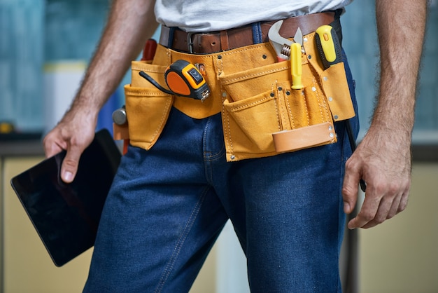 Cropped shot of young repairman wearing a tool belt with various tools holding digital tablet while