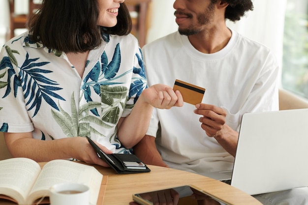 Cropped shot of young woman passing credit card to her husband with laptop while both choosing goods in online shop and paying for them