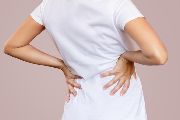 Cropped shot of young woman in a white tshirt holding her lower\
back with her hands on beige color