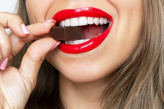 어두운 초콜릿 사탕을 먹고 즐기는 광택 있는 붉은 입술을 가진 젊은 여성의 자른 샷