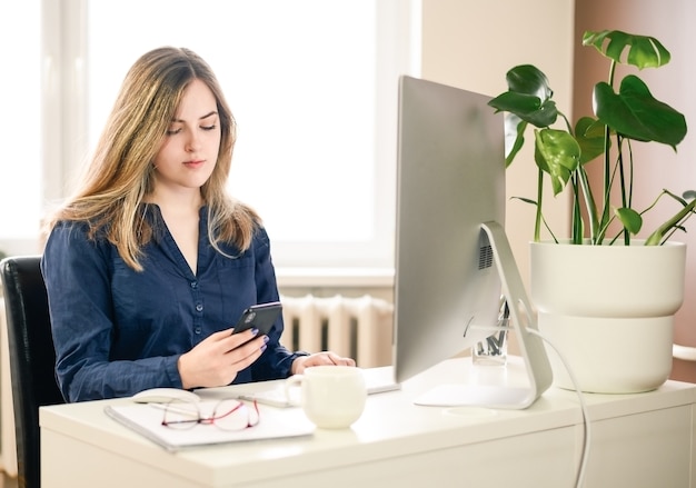 Cropped shot of a young woman working from home using smart phone and computer, woman's hands using smart phone in interior, woman at home workplace using technology,. High quality photo