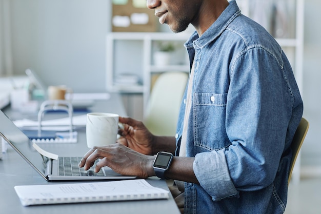 Cropped side view shot of young black man using laptop while\
working in office and holding cup of co