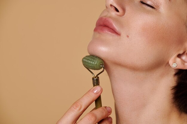 Cropped studio shot of natural beauty caucasian woman with\
fresh clean healthy skin doing face and neck massage using jade\
roller massager. close-up. copy ad space. cosmetology and beauty\
care concept