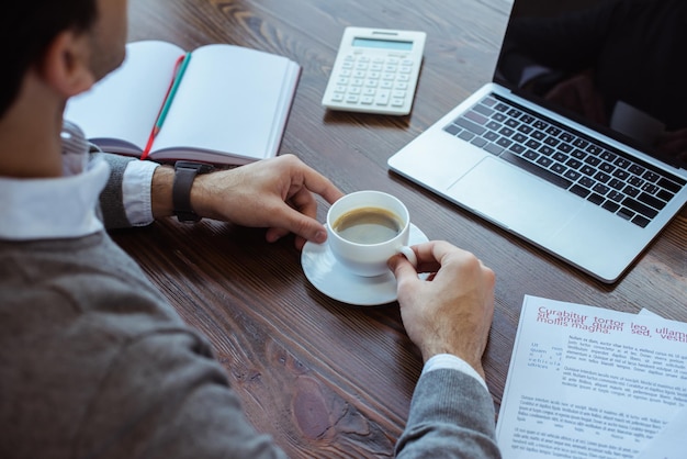 Обрезанный вид бизнесмена с чашкой кофе рядом с бумагами калькулятора ноутбука и блокнотом за столом
