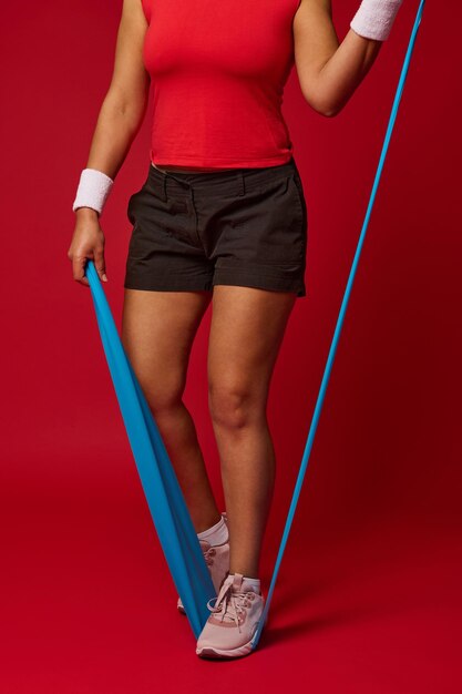 Cropped view of determined fit woman doing body weight workout using elastic strap pulling with arms for shoulder training against red background with copy ad space