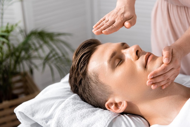 Cropped view of healer standing near man with closed eyes and holding hands above his head