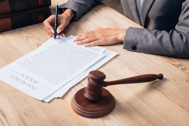 Cropped view of judge signing insurance policy document near gavel in office