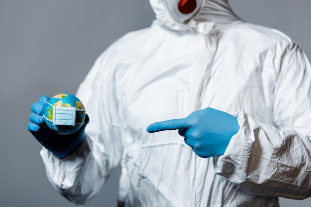 Cropped view of man in hazmat suit pointing with finger at small globe in medical mask isolated on