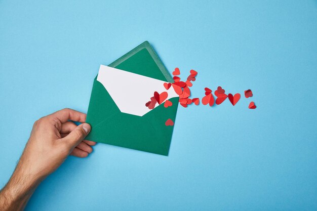 Cropped view of man holding green envelope with blank white card and paper cut red hearts on blue