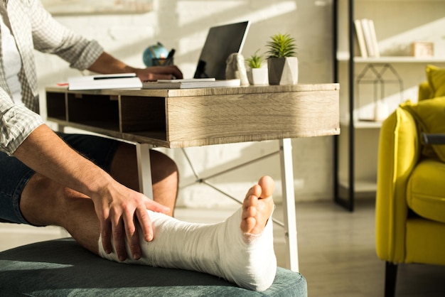 Cropped view of man touching broken leg while using laptop at home