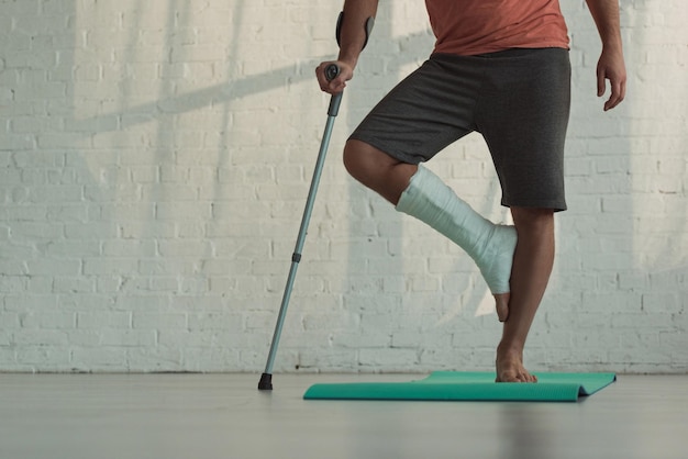 Cropped view of man with broken leg standing on fitness man and holding crutch