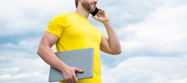 空を背景にスマートフォンで話しているラップトップを持つ男のクロップド ビュー