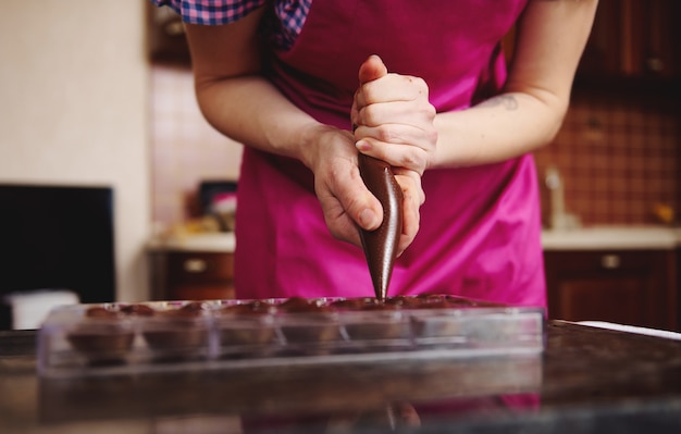 ピンクのエプロンのショコラティエのトリミングされたビューチョコレートの詰め物をチョコレートの型に注ぎ、豪華な手作りのプラリネを準備します