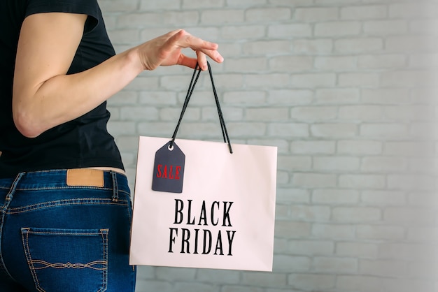 Cropped view sexy woman in denim holding paper bag with tag in her hand against a white brick wall in the mall. Copy space. Black Friday sale concept.