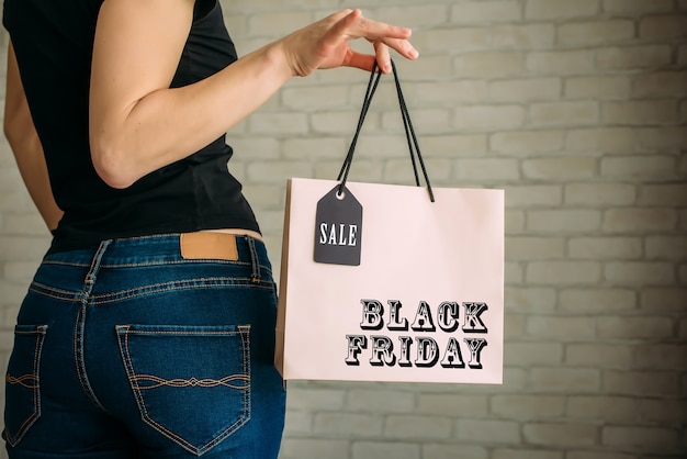 Cropped view sexy woman in denim holding paper bag with tag in her hand against a white brick wall