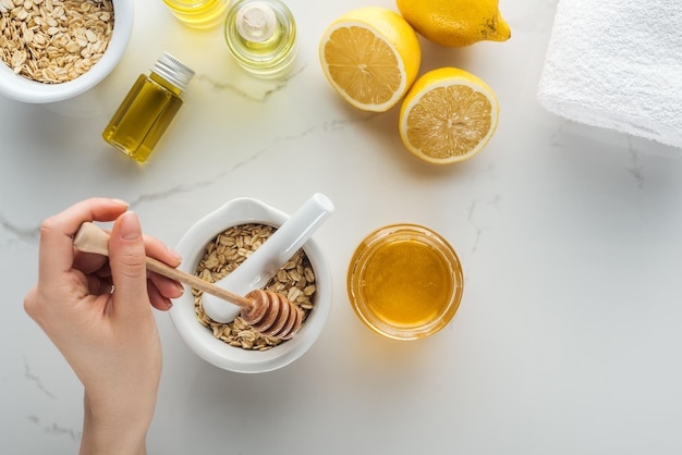 Cropped view of woman adding honey into pounder with oat flakes on white surface