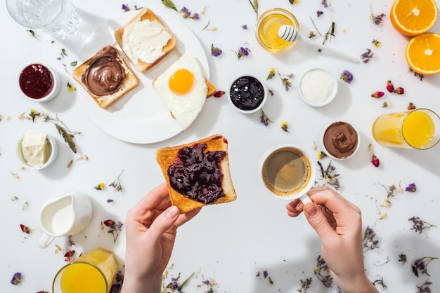 Cropped view of woman holding cup with coffee and toast with jam on white