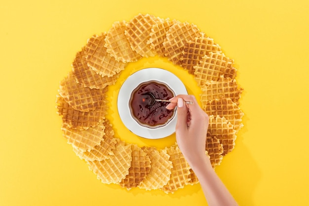 Cropped view of woman holding teapot in cup of tea rounded by waffles on yellow