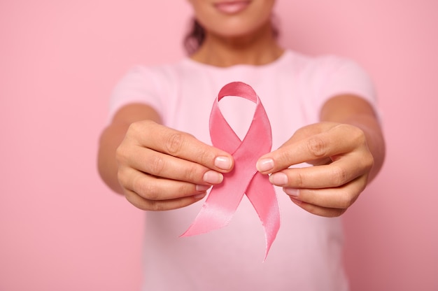 Cropped view of a young woman in pink t-shirt holding a pink\
satin ribbon isolated on colored background with copy space.\
international breast cancer awareness day, breast cancer support\
concept.