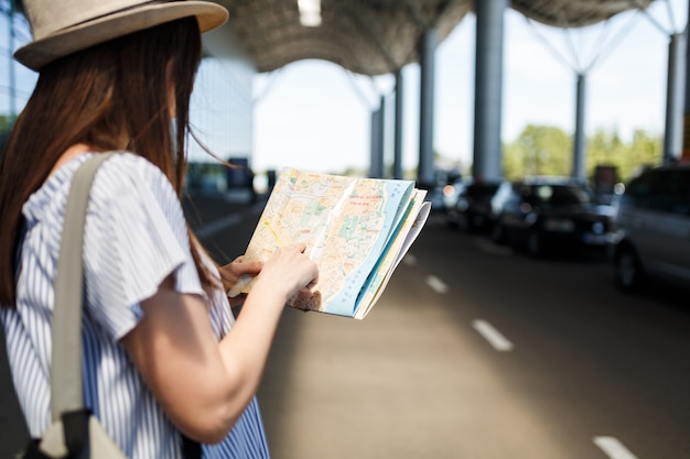 国際空港で紙の地図のバックパック検索ルートと帽子をかぶった若い旅行者観光客の女性をトリミング