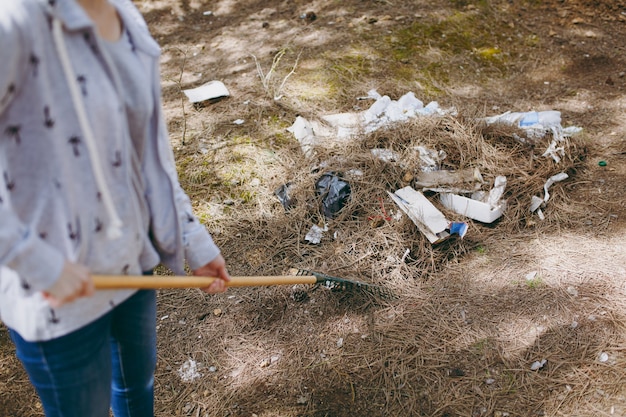 Foto giovane donna ritagliata in abiti casual che pulisce la spazzatura usando il rastrello per la raccolta dei rifiuti nel parco disseminato