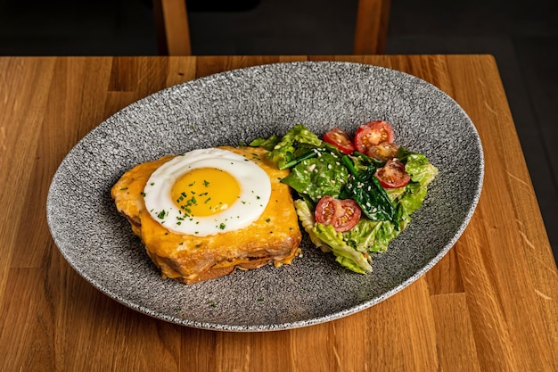 Croque madame breakfast served on a grey plate with salad mix and cherry tomatos for garnish on a wooden table in a restaurant