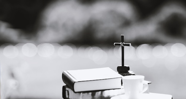 The cross of Jesus Christ, the Bible and a teacup on the shore of a serene lake in the sunlight