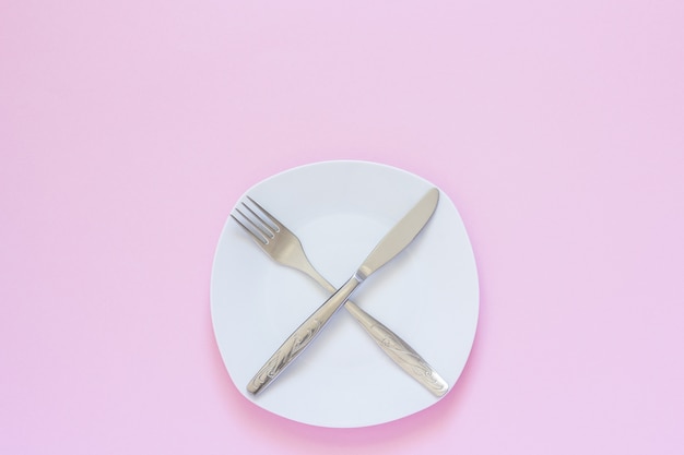 Crossed fork and knife on white plate on pink background.